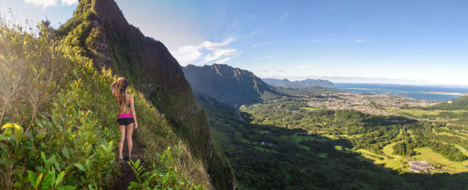 A hiker on the Nuuanu Saddle ridge trail on Oahu, overlooking lush valleys, jagged mountains, and the Pacific Ocean in the distance.