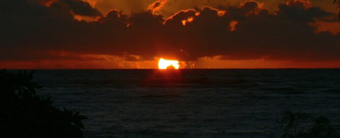 A vibrant Hawaiian sunrise over the ocean, with golden light reflecting on the waves and clouds glowing in deep orange hues.