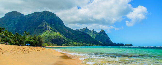 Golden sand, turquoise ocean, and lush green mountains at Ha'ena Beach on Kauai’s North Shore under a partly cloudy sky.