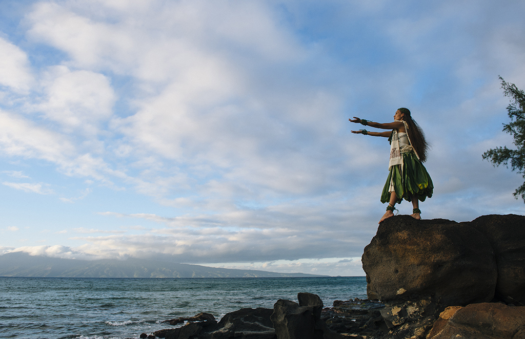 Woman hula dancing on top of coastal rocks wearing traditional clothing