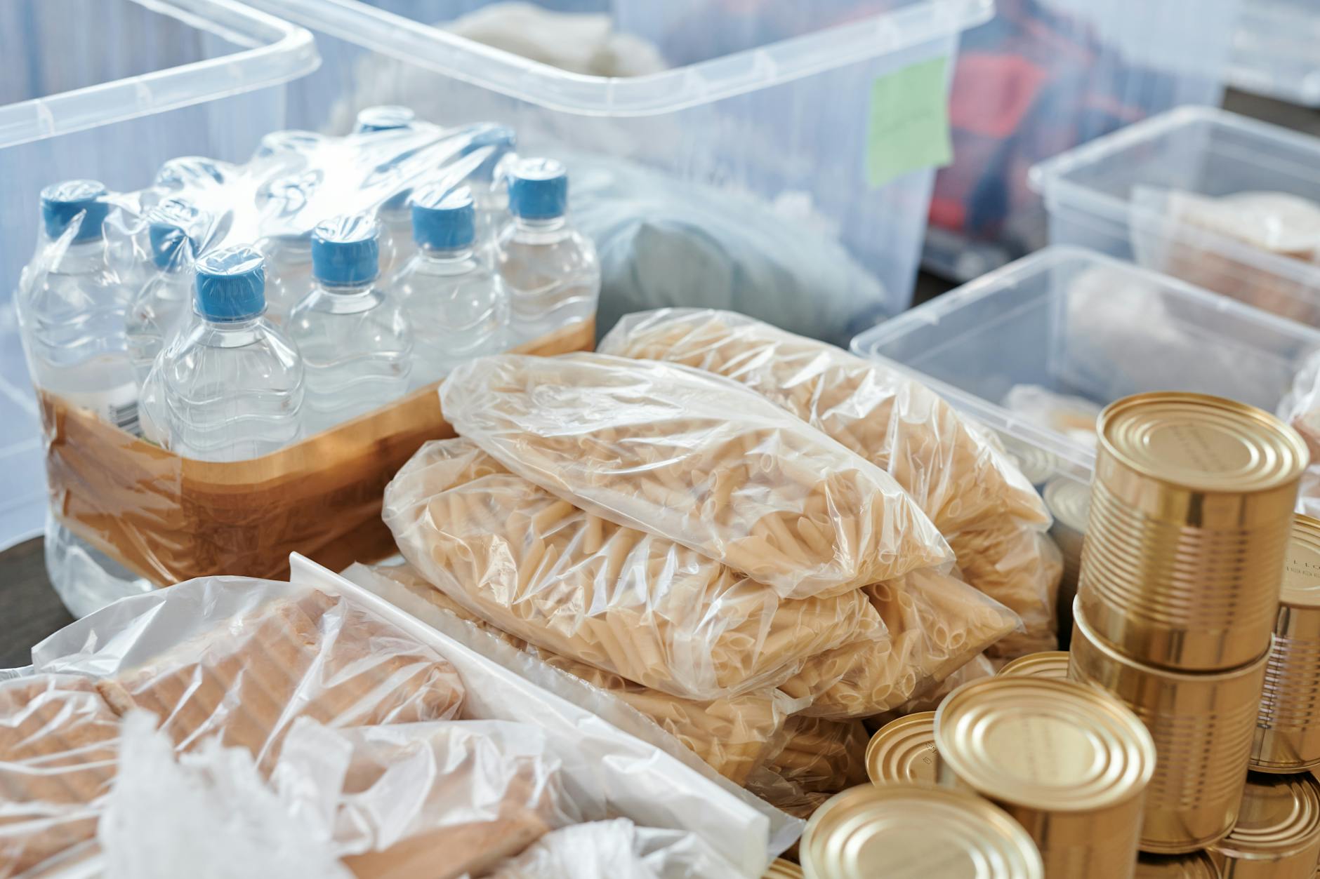 Plastic-wrapped food items and bottled water organized in storage containers and tin cans. (Photo by Julia M Cameron via Pexels)